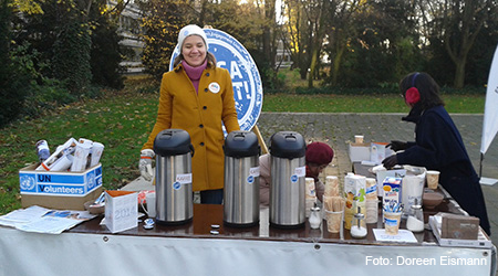 Junge Frau hinter Tisch mit Kaffeekannen und Geschirr, im Freien; das Logo der UN Volunteers und Teile einer Werbetafel mit einem überdimensionierten „Engagiert“-Anstecker, der eine Kampagne von Engagement Global begleitet. Foto: Doreen Eismann.