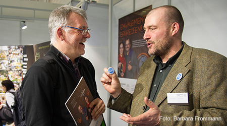 Zwei Männer unterhalten sich angeregt auf dem Stand von Engagement Global auf der Fachmesse didacta. Im Hintergrund ein zielgruppenspezifisches Plakat der Werbekampagne von Engagement Global. Foto: Barbara Frommann.