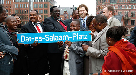 Sieben Vertreter und vier Vertreterinnen afrikanischer Kommunen zeigen ein Schild mit der Aufschrift: Dar-es-Salaam-Platz. Im Hintergrund sind Kontorhäuser der historischen Speicherstadt zu sehen. Foto: Andreas Grasser.