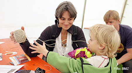 Junge Frau sitzt an einem Tisch mit Broschüren und hält ein Knäuel mit Paketschnur, an der Schnur zieht ein kleines Kind. Im Hintergrund ein junger Mann, der die beiden beobachtet. Foto: BMZ.